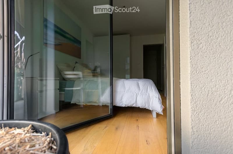 Modern bedroom with a large glass sliding door leading to a balcony, featuring a neatly made bed with white bedding, a nightstand with a lamp, and a mirror on the wall.
