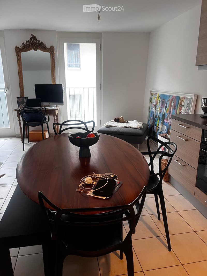 A well-lit room with a round wooden table surrounded by chairs, a screen on a desk, a mirror, a stool, and a colorful picture on the wall.