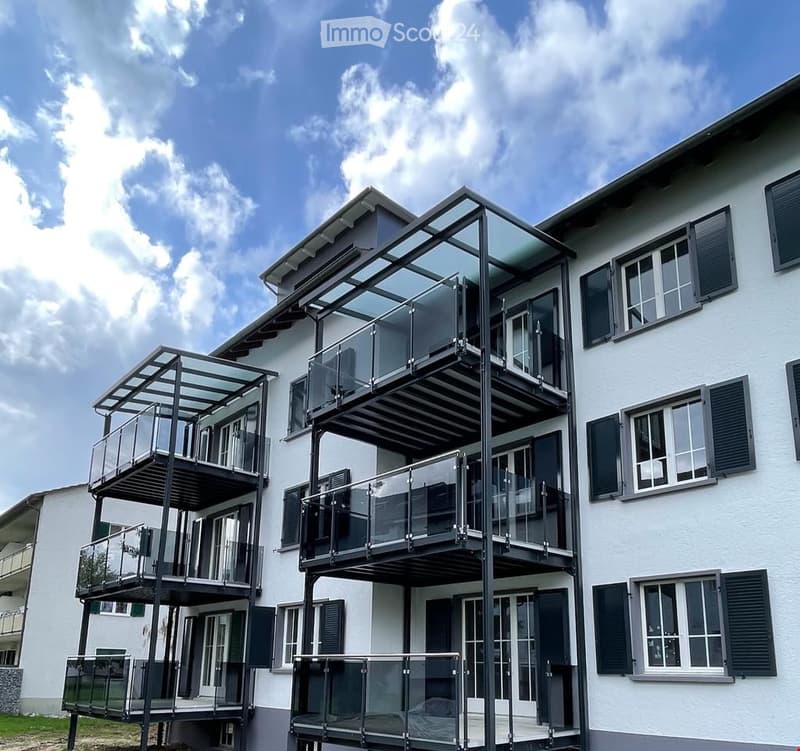 Modern two-story building with white exterior, multiple large windows with shutters, and several glass-enclosed balconies.