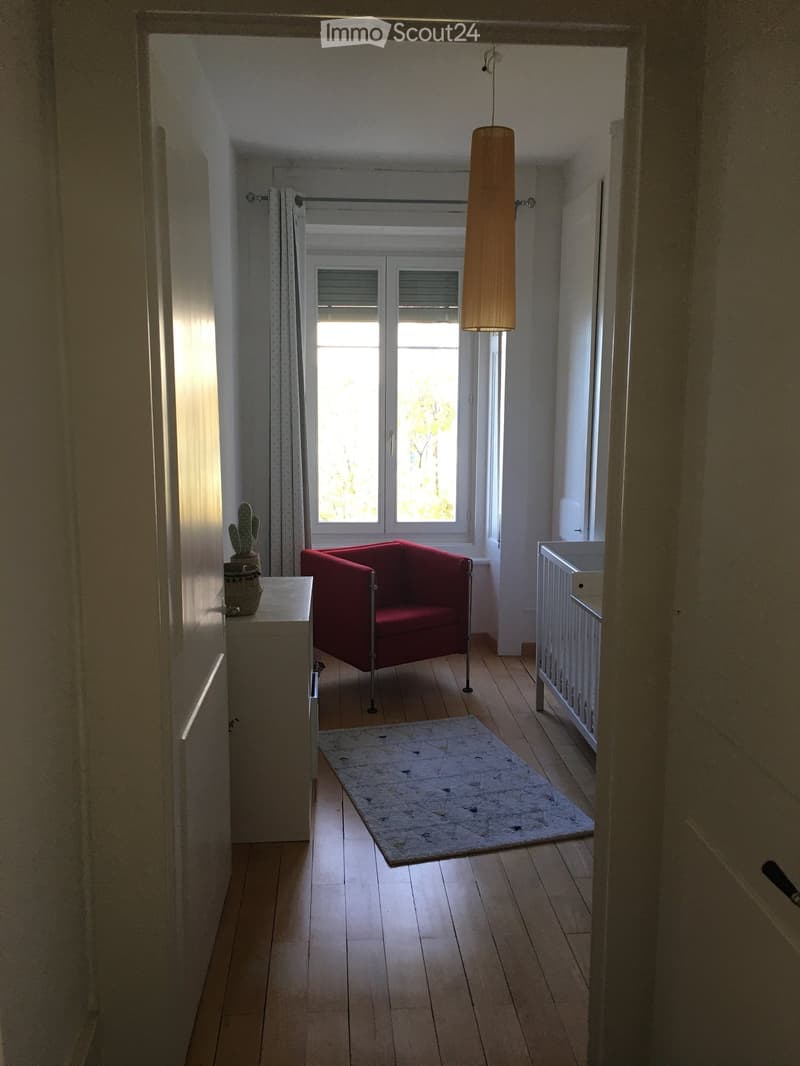 A narrow hallway with wooden flooring, a red sofa, a small table with a cactus, a baby crib, and a patterned rug.