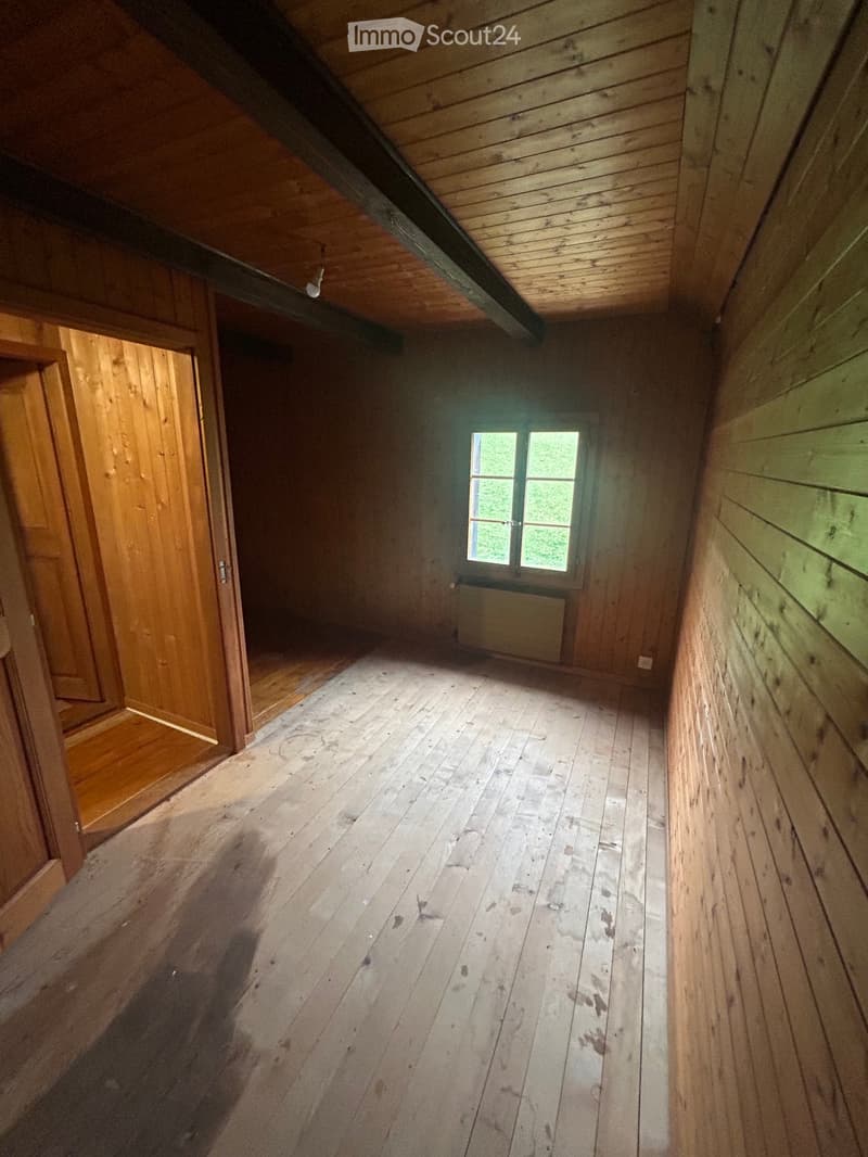 Empty wooden room with exposed beams, wooden floor, single window, and a door leading to another room.