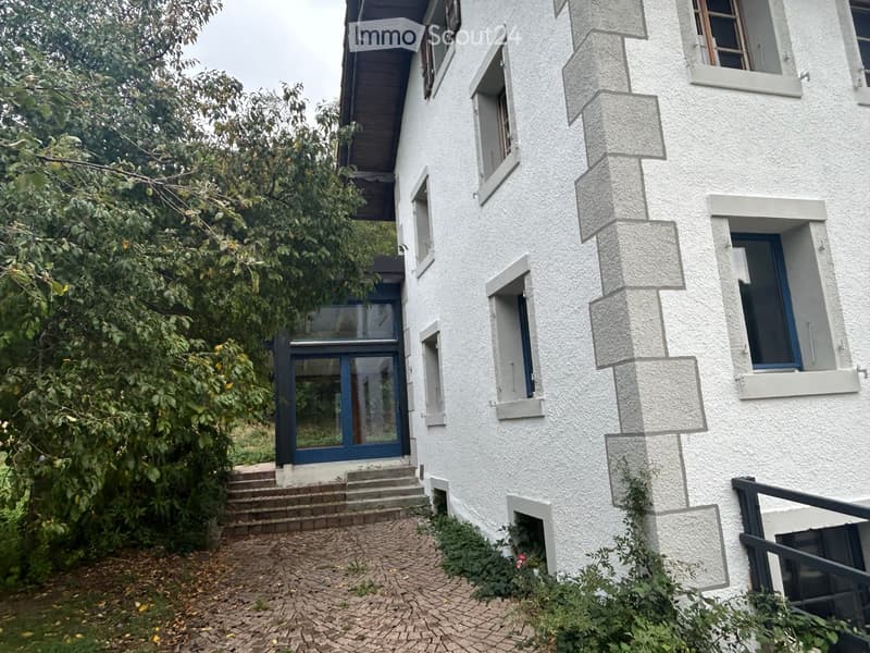 Two-story white house with multiple windows, a raised ground floor, and a small garden area.