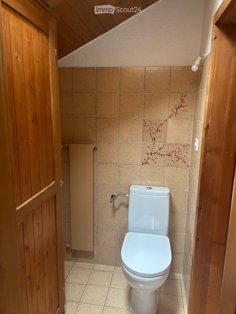 A small bathroom with beige tiled walls and floor, featuring a white toilet and a wooden cabinet.