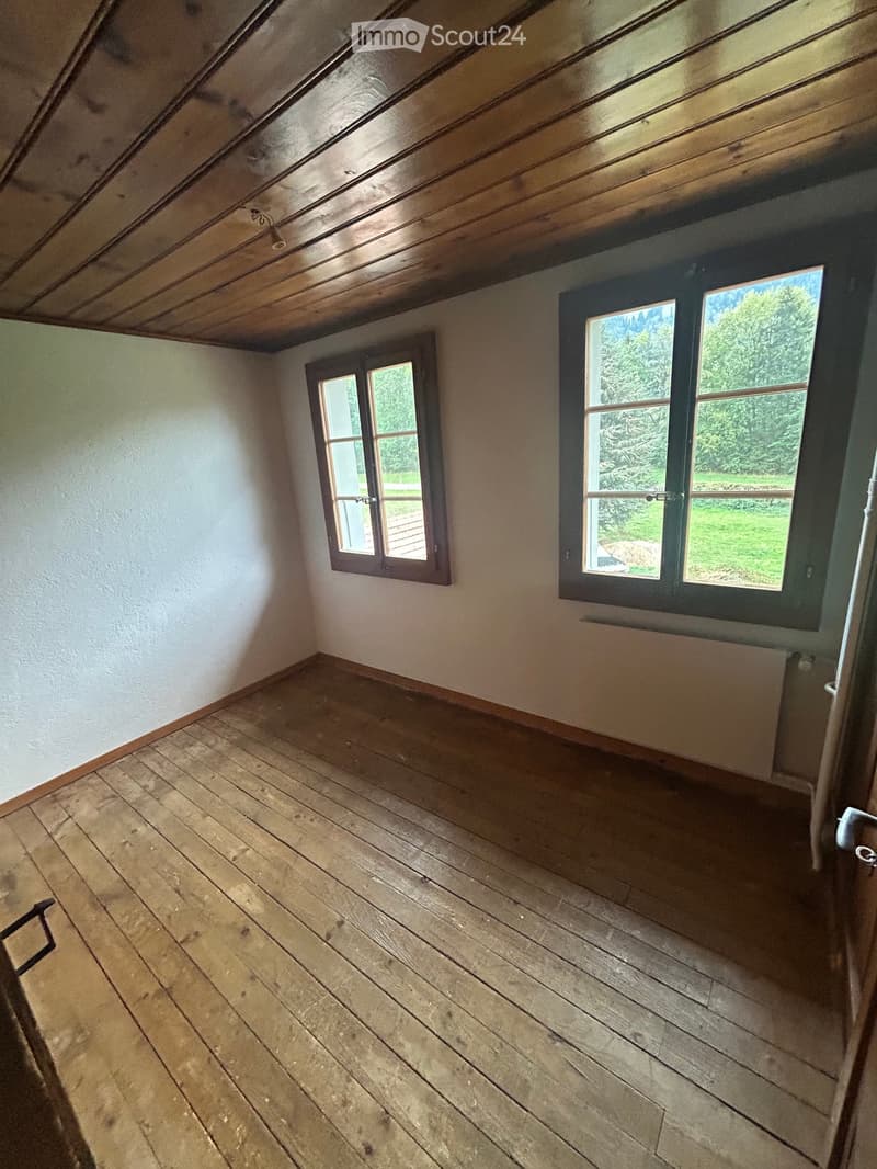 Empty attic room with wooden floors, exposed wooden ceiling beams, and two windows offering a view of greenery outside.