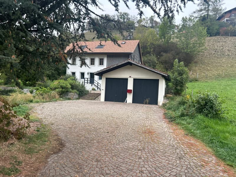 Two-story house with a red-tiled roof, white exterior, and a two-car garage. The house is surrounded by greenery and trees, with a cobblestone driveway leading to the garage.