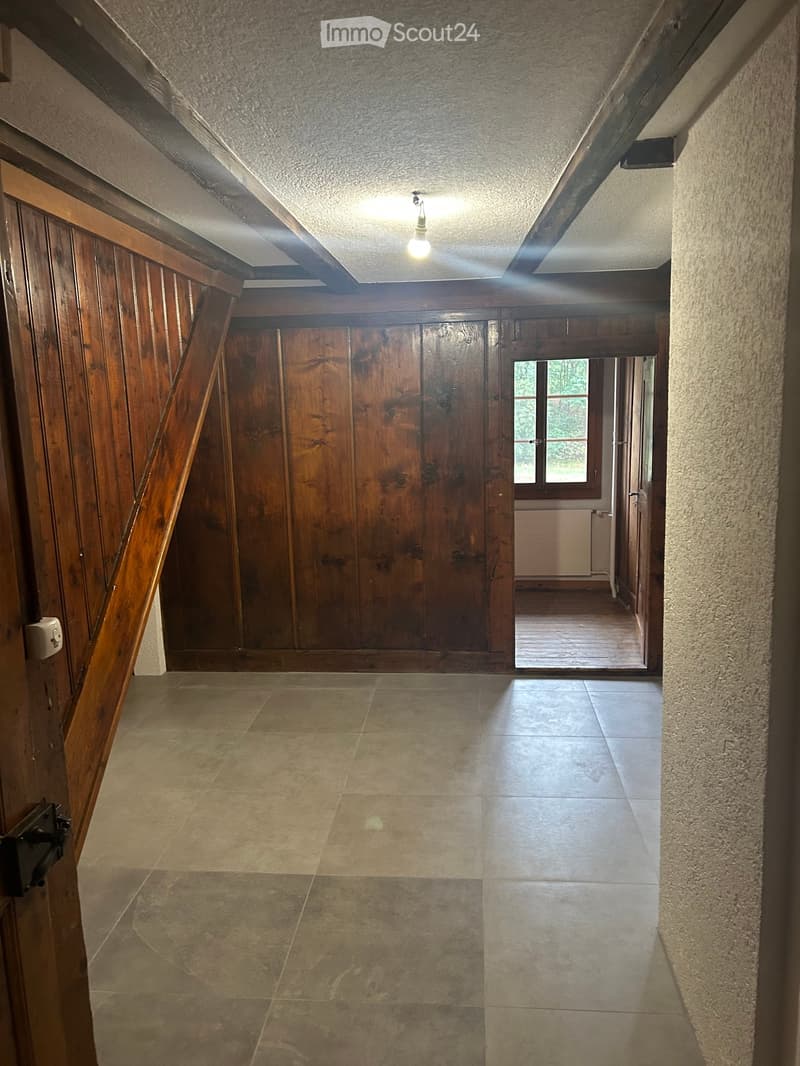 Empty hallway with exposed wooden beams, tiled floor, and a window at the end leading to another room.