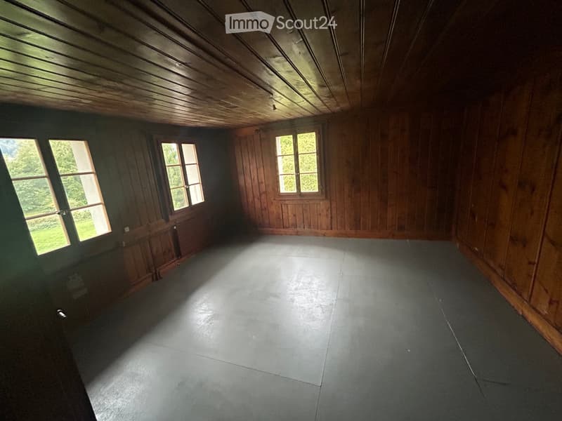 Empty room with wooden walls, wooden ceiling, and concrete floor. Three windows with wooden frames and glass panes, allowing natural light to enter. The room is completely unfurnished with no visible furniture or equipment.