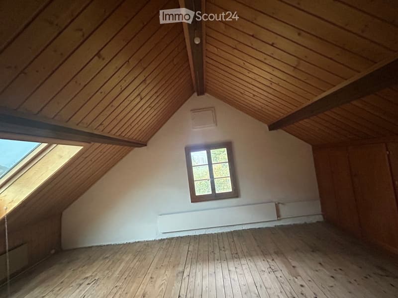Empty attic room with wooden slanted ceilings, wooden floor, and a single window with a brown frame. The walls are white and the room is well-lit with natural light coming through the window and skylight.