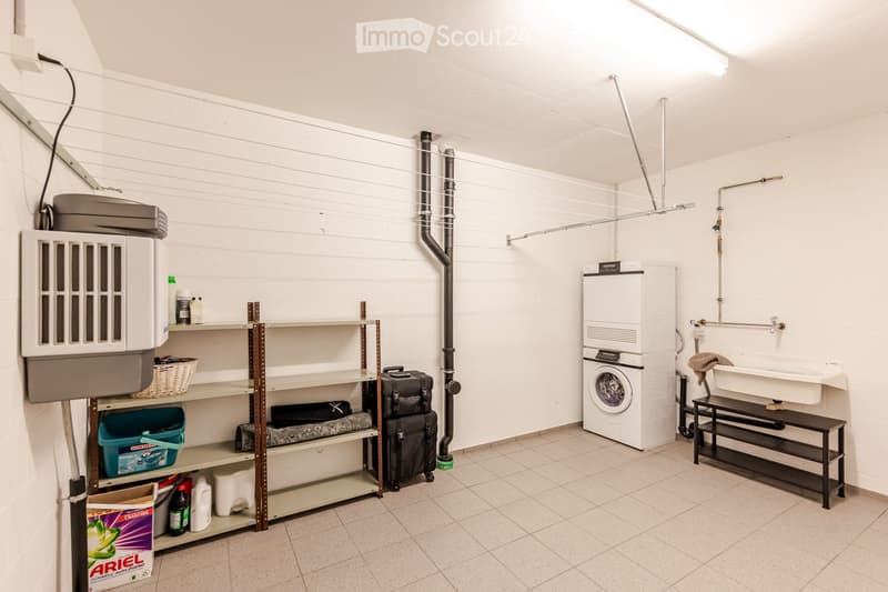 Utility room with a washing machine, sink, shelves, and various stored items including a suitcase and cleaning supplies.