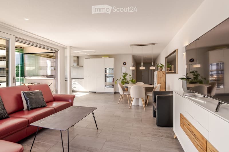 Modern living room with a red leather sofa, coffee table, dining area with chairs, and a television. The space features a balcony with large glass doors, white cabinets, and a sleek design.