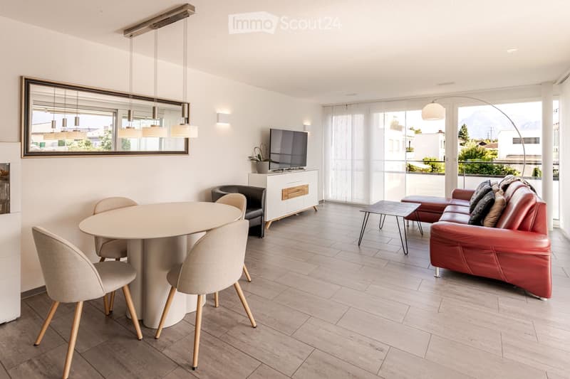 Modern living room with a red leather sofa, round dining table with four chairs, TV stand, and large windows offering a scenic view.