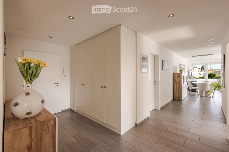 A modern hallway with wooden flooring, white walls, and built-in wardrobes. A large white vase with yellow and white flowers sits on a wooden console table. The hallway leads to a dining area with white chairs and a table.