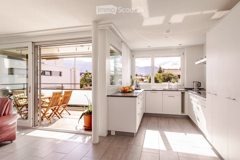 Modern kitchen with white cabinets, a central island, and a dining area with wooden chairs and a table. The space features large windows offering a scenic view of the mountains.
