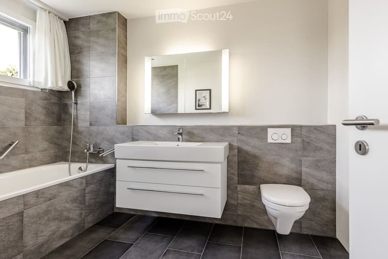 Modern bathroom with a bathtub, sink, toilet, and a large mirror above the sink. The walls are tiled in gray, and the floor is dark tiled.