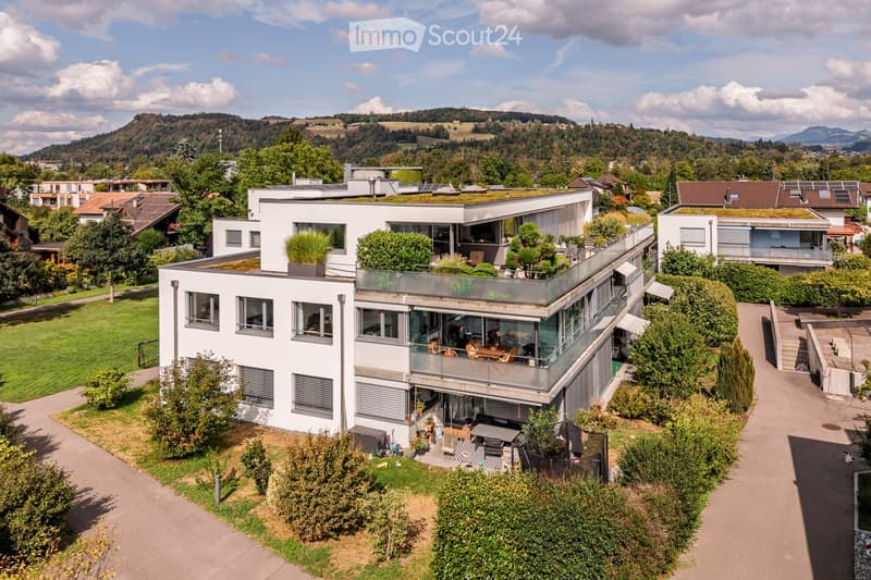 Modern white house with large windows, green roof, and surrounding garden with outdoor seating area.