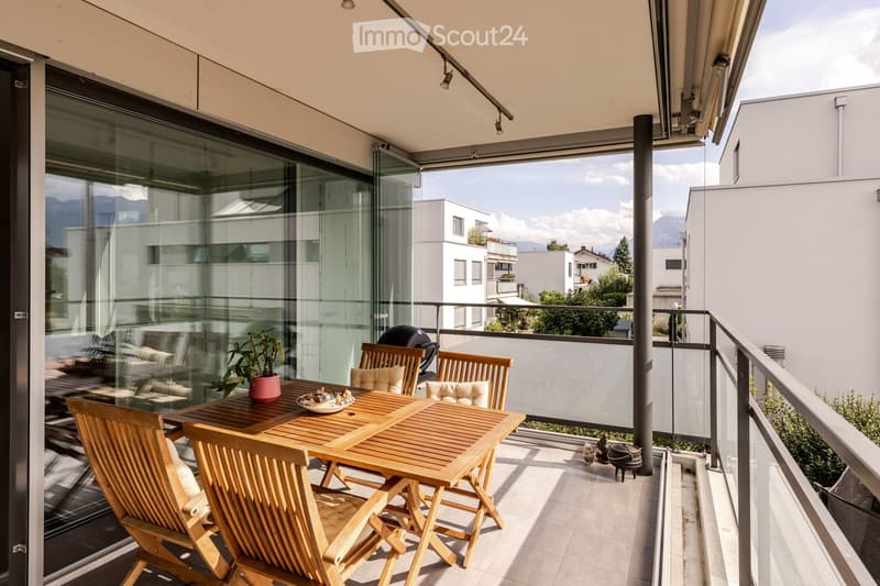 Balcony with wooden table, chairs, potted plant, and grill, overlooking a cityscape with mountains in the background.