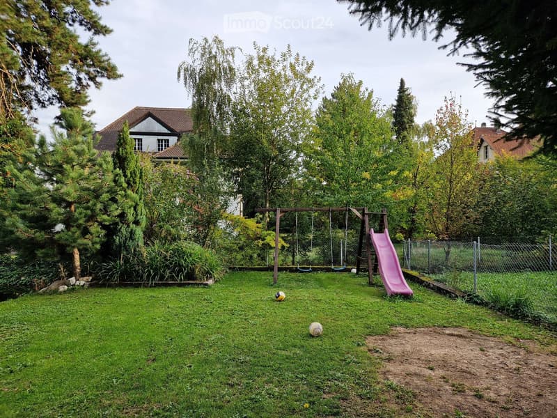 A well-maintained garden with a swing set, slide, and two balls on the grass, surrounded by trees and a house in the background.
