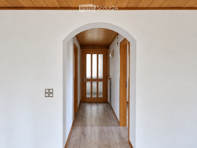 A modern hallway with light wooden flooring, white walls, and a wooden door at the end. The ceiling is also wooden, and there are light switches and a wall-mounted light fixture visible.