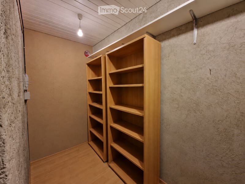 A small, well-lit room with wooden flooring and two tall, empty wooden shelves against the wall. The walls are textured, and the ceiling has recessed lighting.