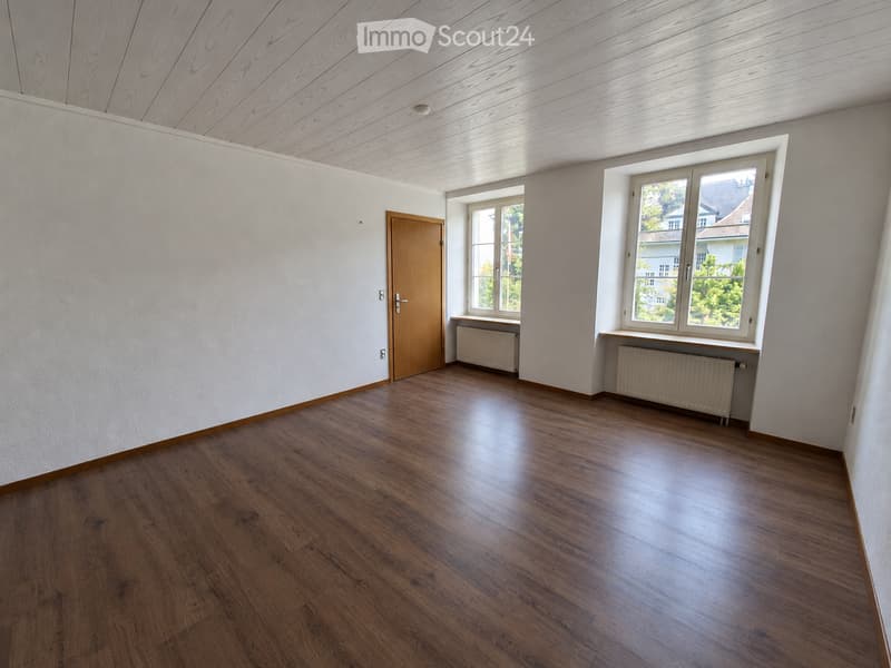 Empty room with wooden flooring, white walls, and large windows offering a view of the outside. The room features a wooden door and radiators under the windows.