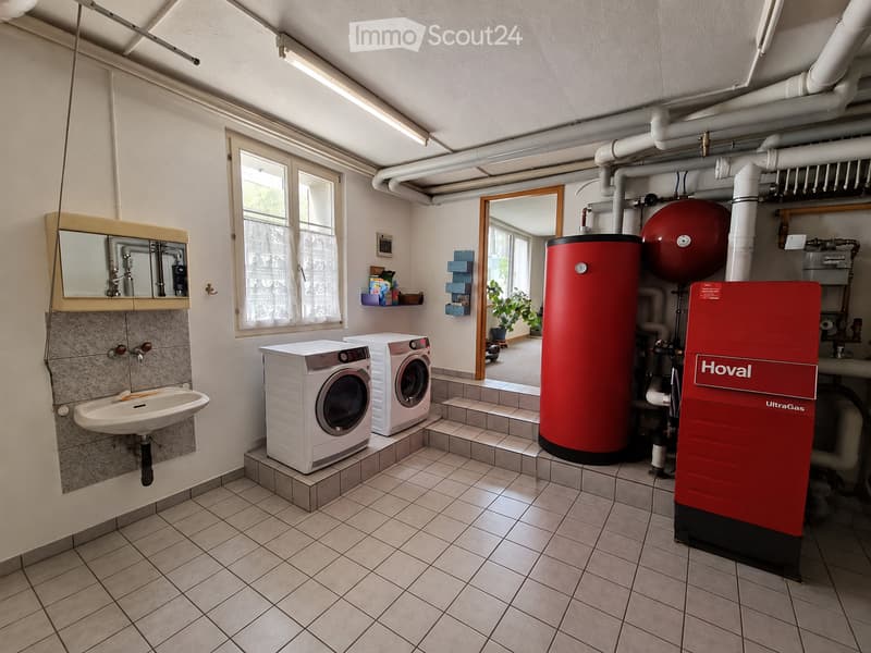 Bathroom with a sink, mirror, washing machine, dishwasher, and water heater. The room has tiled flooring and walls, with a window and shelves holding various items.