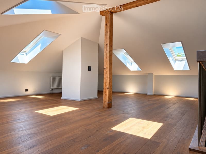 Bright, empty attic space with wooden flooring, white walls, and multiple skylights allowing natural light to flood the room. A single wooden support beam is visible in the center.