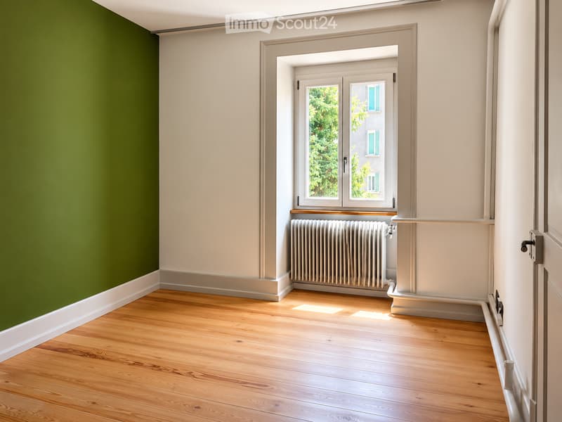 Empty room with wooden flooring, light-colored walls, green accent wall, window with radiator below, and door visible on the right side.