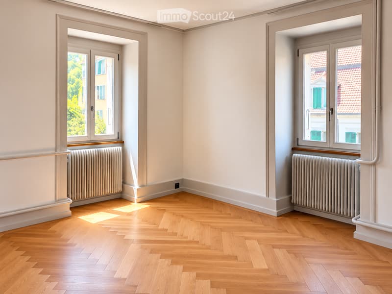 Empty room with light wooden flooring, white walls, and two windows with radiators beneath them.