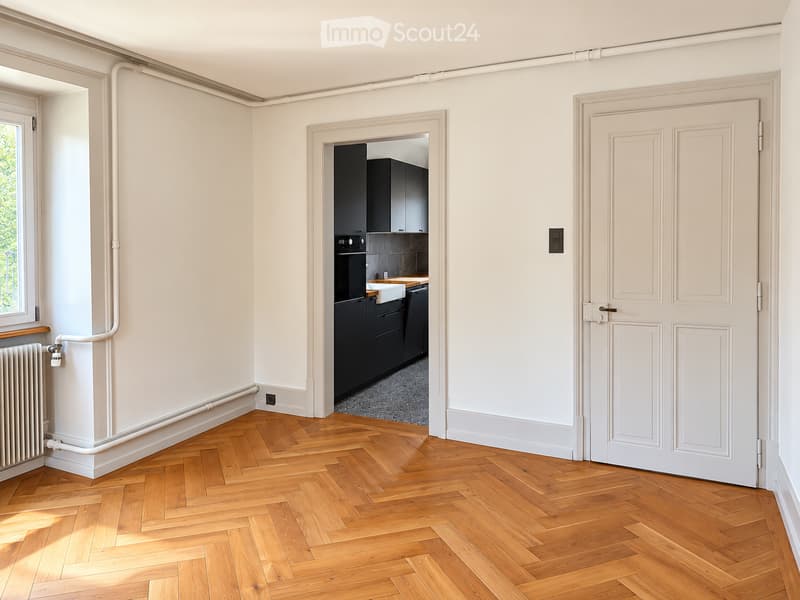 Empty room with light-colored walls, wooden herringbone flooring, and a white door leading to a modern kitchen area with black cabinets and stainless steel appliances.