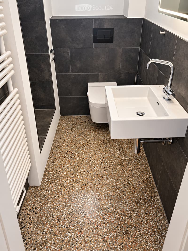 Modern bathroom with dark grey tiled walls, a white sink with a chrome faucet, and a white toilet. The floor features a colorful, speckled mosaic tile design.