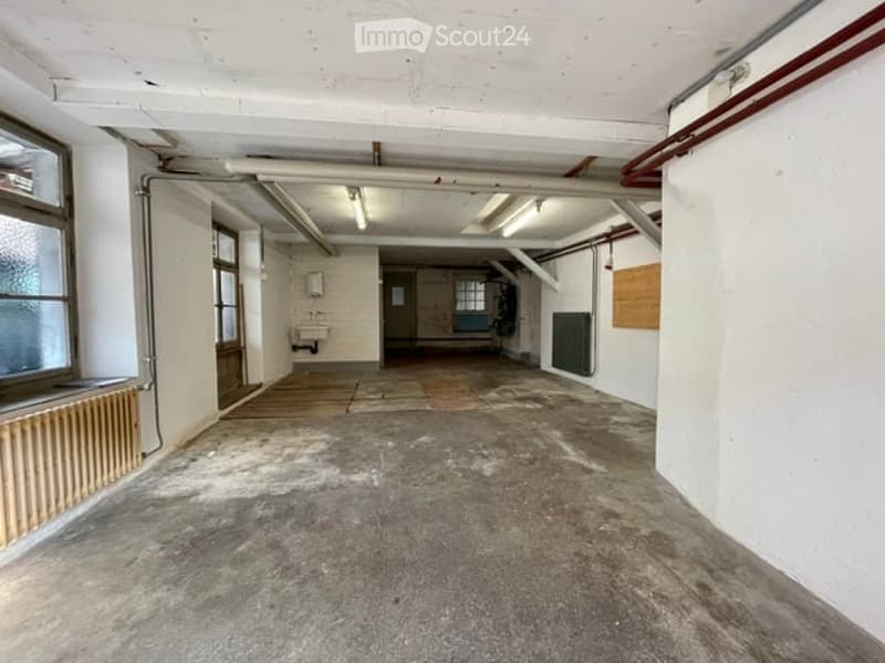 Empty industrial-style room with concrete floor, exposed pipes, and large windows. No furniture or equipment present.