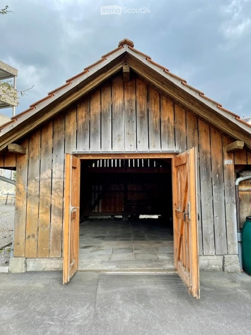 A rustic wooden barn with a triangular roof and open double doors, situated on a paved area.