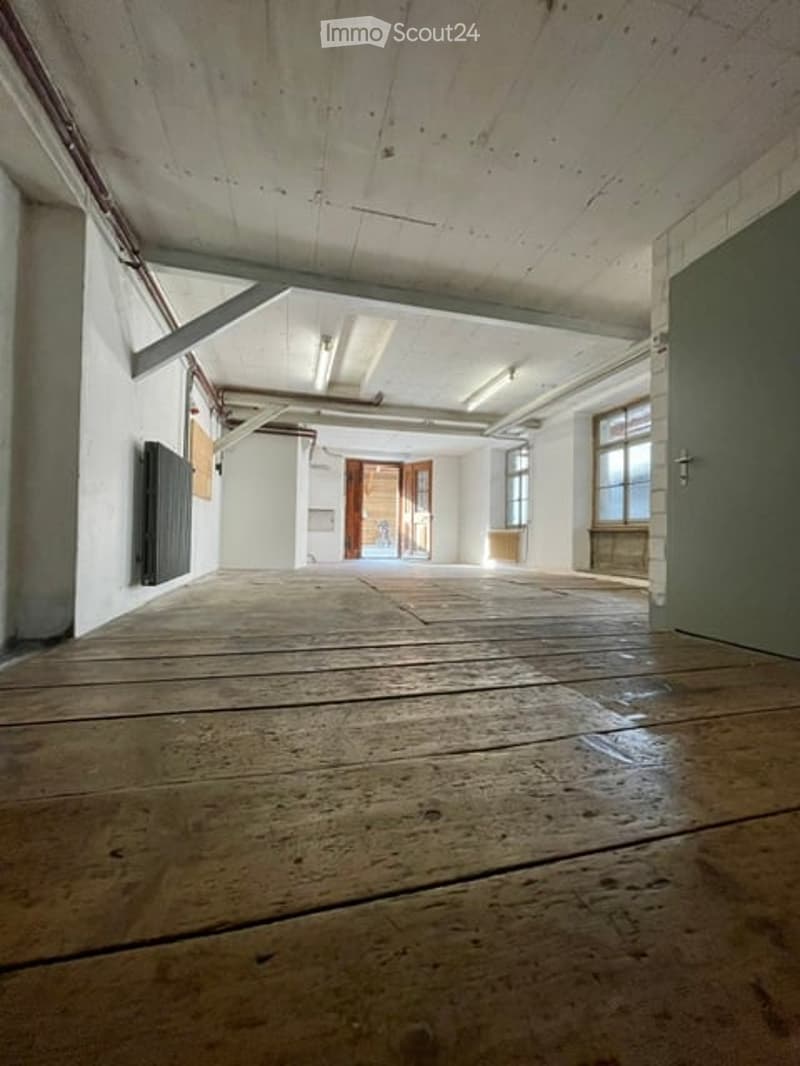 Empty room with wooden floor, white walls, exposed beams, and large windows. No furniture or equipment present.