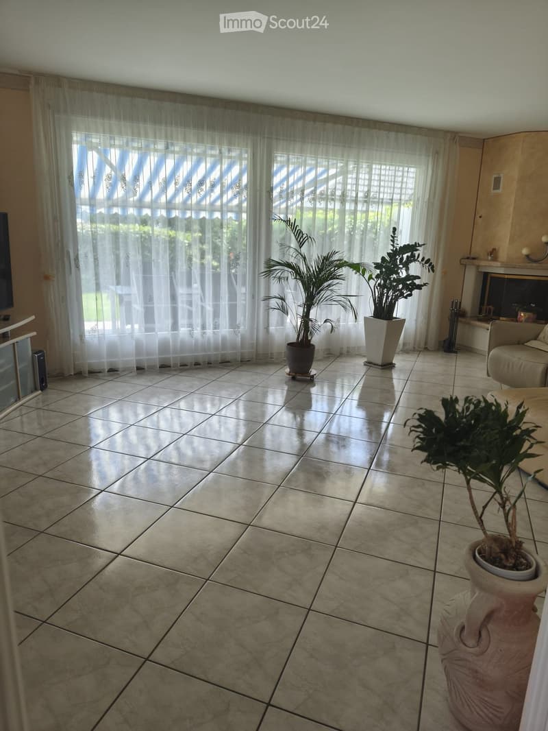 Bright living room with tiled floor, large windows covered by sheer curtains, a beige sofa, a television on a stand, and several potted plants.