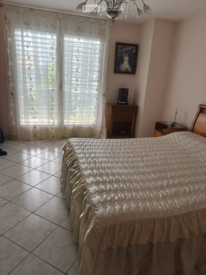 A well-lit bedroom with a large bed covered in a beige quilt, a nightstand with a lamp, a wooden drawer unit, and a framed picture on the wall. The room features tiled flooring and sheer curtains on the windows.