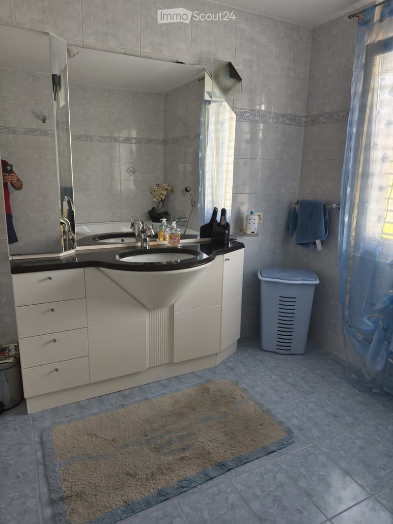 Bathroom with double sink, mirror, white cabinets, tiled walls and floor, towel, trash bin, and a small rug.