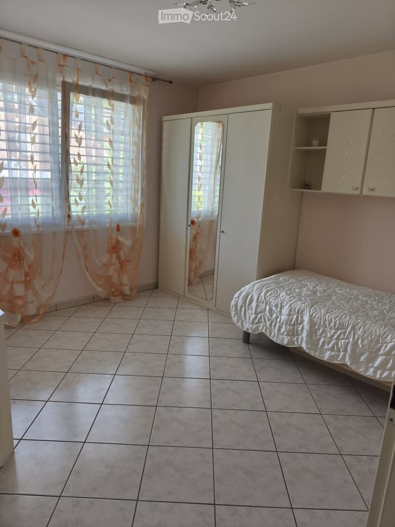 A small bedroom with tiled flooring, a bed with a white quilt, a large wardrobe with mirrored doors, and a window with orange curtains.