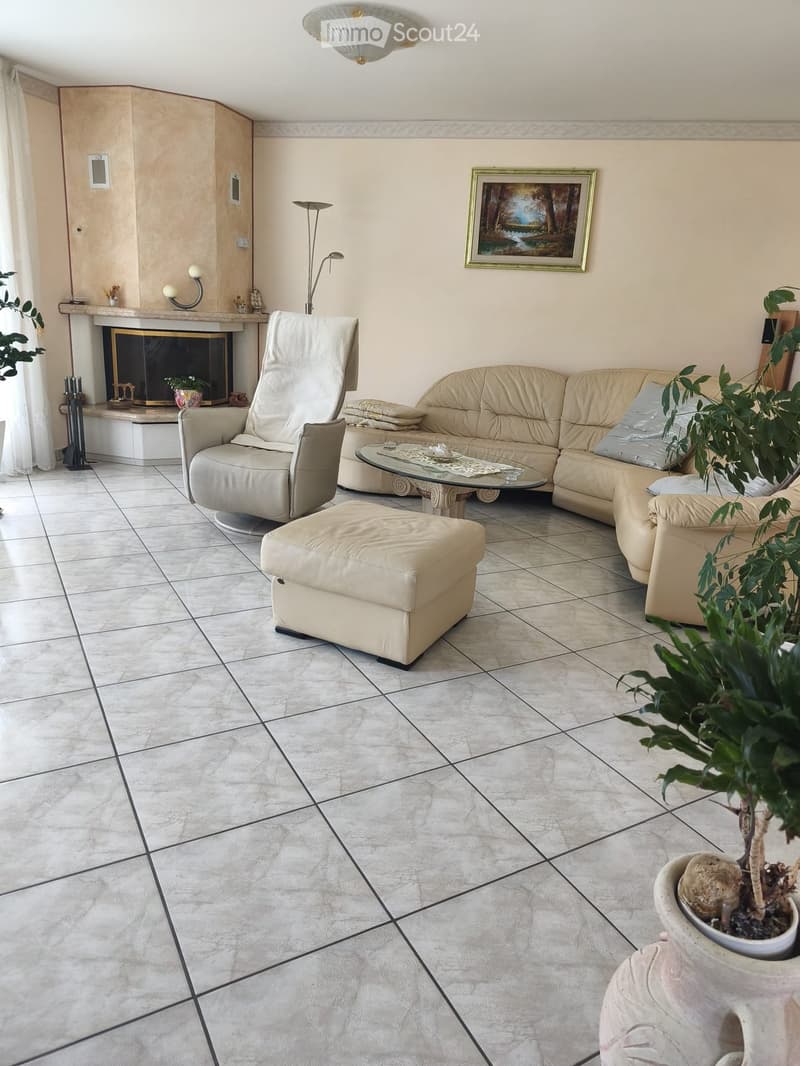 Living room with beige sofa, glass coffee table, ottoman, armchair, fireplace, and decorative plants.