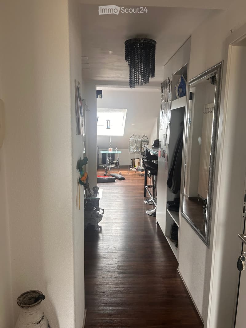 A long, narrow hallway with wooden flooring, white walls, and a chandelier hanging from the ceiling. The hallway leads to a room with a small table, chairs, and a window letting in natural light.