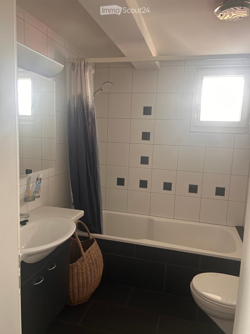 Modern bathroom with white tiles, a bathtub, a sink, a toilet, a mirror, and a shower curtain. A wicker basket is placed on the floor near the sink.