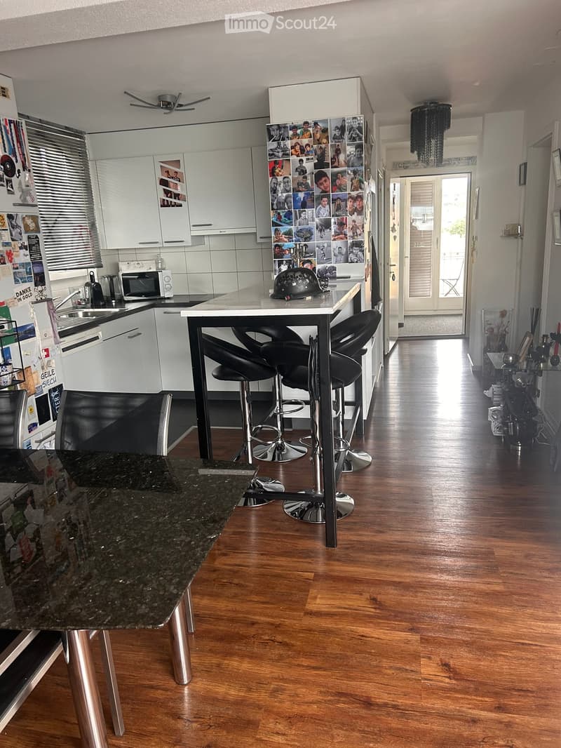 Modern kitchen with white cabinets, granite countertops, and a central island with four black bar stools. The kitchen features a stainless steel sink, microwave, and refrigerator, with a hallway leading to another room.