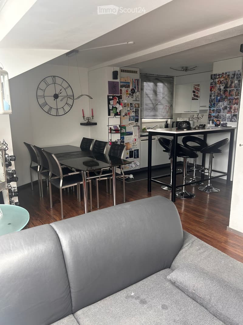 Modern open-plan living and dining area with a grey sofa, dining table with chairs, bar table with stools, and a kitchen area with cabinets and a refrigerator.