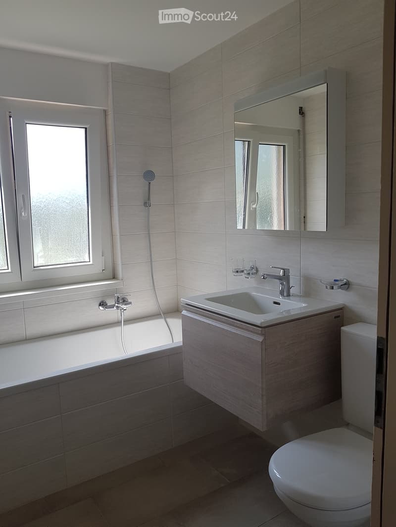 Modern bathroom with a bathtub, sink, mirror, and toilet. Features a large window with frosted glass and a sleek wooden vanity cabinet.