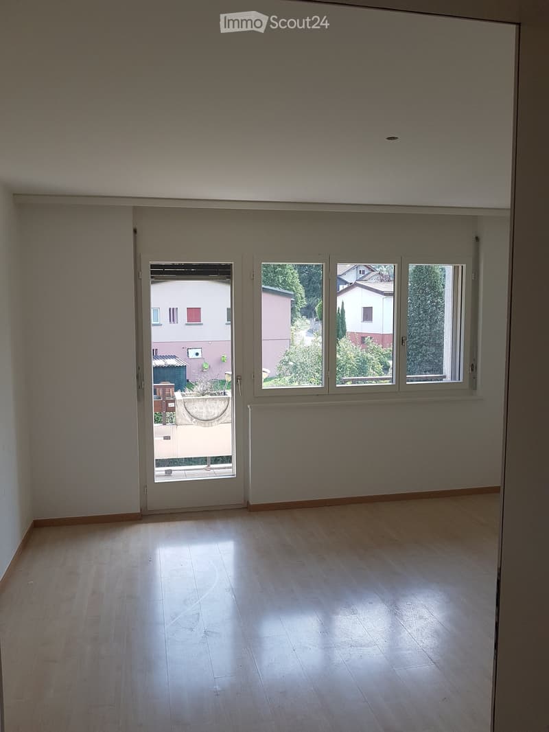 Empty room with wooden flooring, large windows, and a sliding door leading to a balcony or outdoor area.