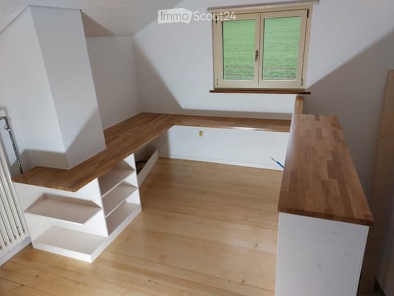 An empty room with wooden flooring, white walls, built-in wooden shelving, and a window with white frame. No furniture or equipment present.