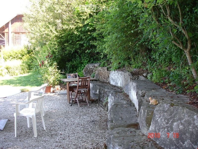 Outdoor seating area with a wooden table, wooden chairs, and white plastic chairs, surrounded by greenery and stone walls.