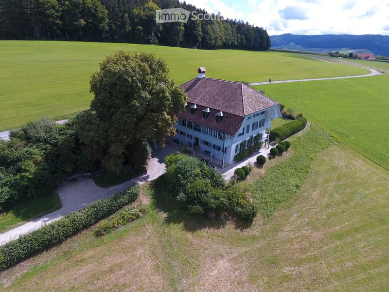 A two-story house with a red-tiled roof and white walls, surrounded by a well-maintained garden and open fields.