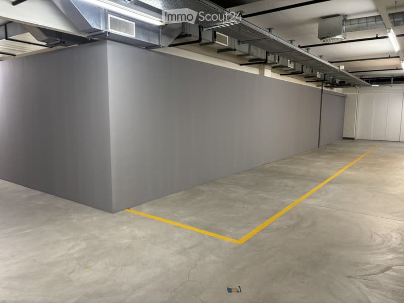 Empty underground parking space with grey concrete walls, concrete floor, yellow parking lines, and exposed ceiling pipes and lighting.