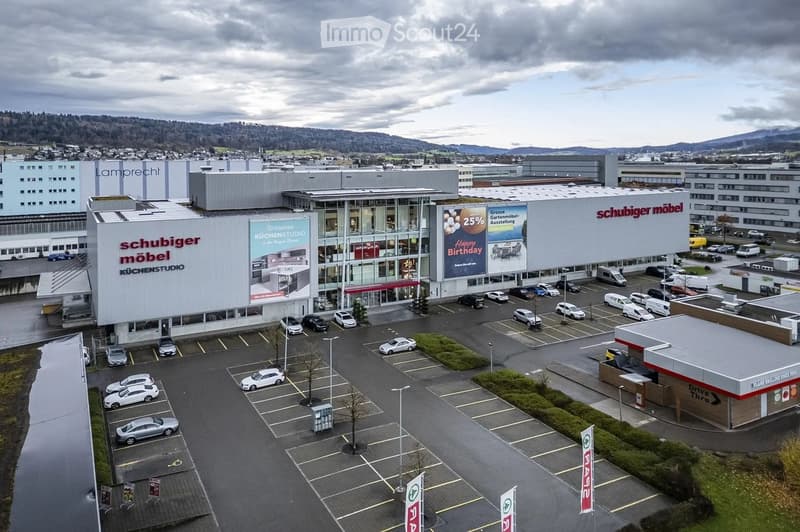 Aerial view of a large commercial building complex with multiple parking lots, several cars, and signage for "Schubiger Möbel" and "Küchenstudio".
