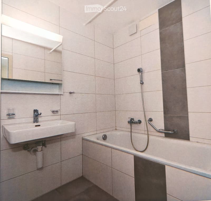 Modern bathroom with a white sink, bathtub, and built-in cabinet with a mirror above it. The walls are tiled in light beige with a dark accent wall.